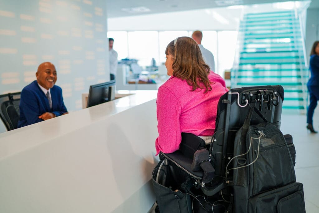 An office receptionist welcomes a visitor who is a wheelchair user