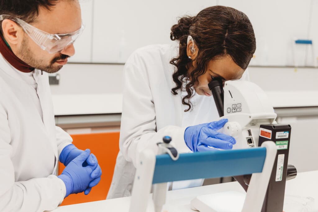 A person who is Deaf and wears a hearing id looks through a microscope beside a colleague .