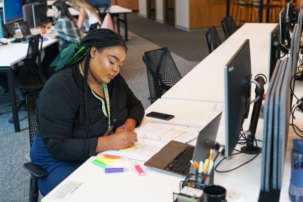 Person wearing sunflower lanyard to show less visible disabilities (such as ADHD, mental health condition, Dyslexia) sits at desk writing on post-it notewith coloured highlighters and a notepad on the desk and an open plan office behind them.