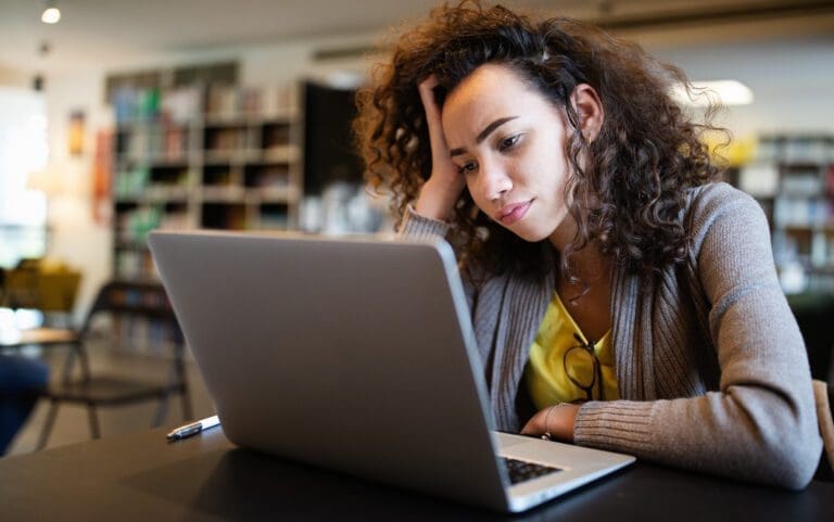 White woman with brown curly hair sitting at a table and working on her laptop. She looks confused while looking at the screen.