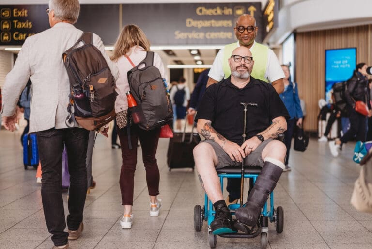 Person wearing a medical boot and using a mobility aid being pushed in a wheelchair by an airport worker.