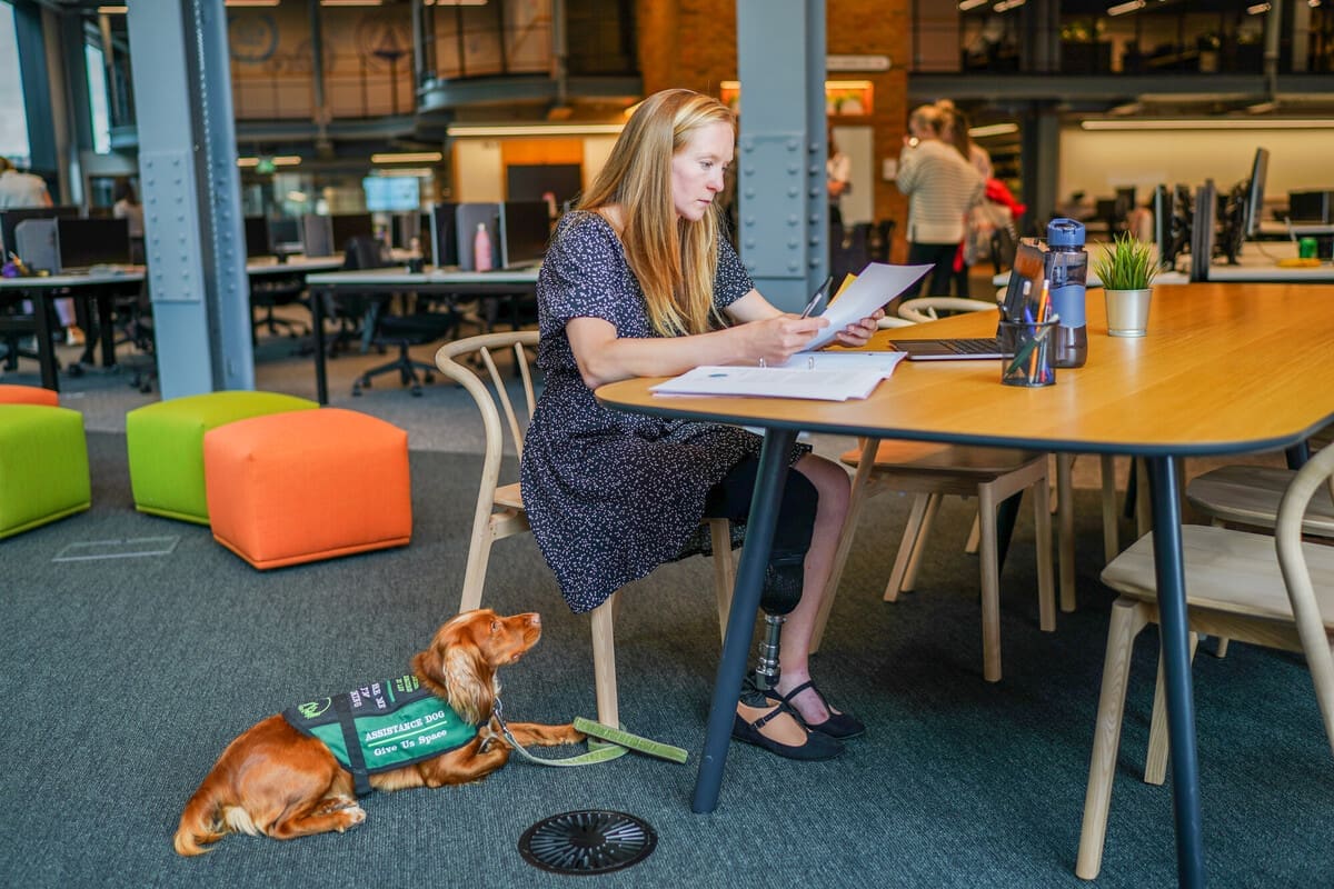 Professional who wears a prosthetic leg works at a desk in an office, with their assistance dog lying at their feet.