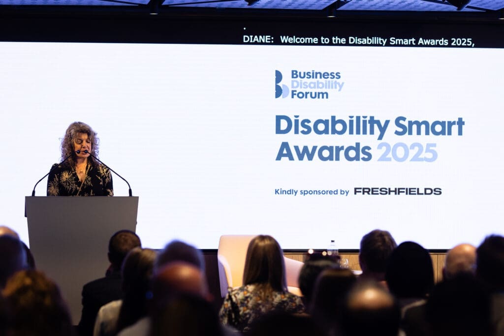 Diane Lightfoot, CEO, Business Disability Forum, hosting the Disability Smart Awards 2025 ceremony onstage. She stands behind a lectern speaking into a microphone. The Awards logo is projected onto a screen behind her.