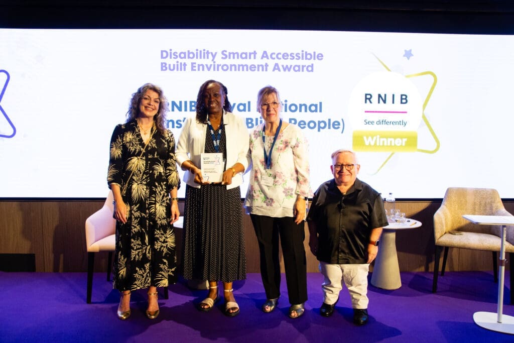 The RNIB team posing on stage with ceremony hosts Diane Lightfoot (left) and Simon Minty (right) after winning the Accessible Built Environment Award at the Disability Smart Awards 2025.