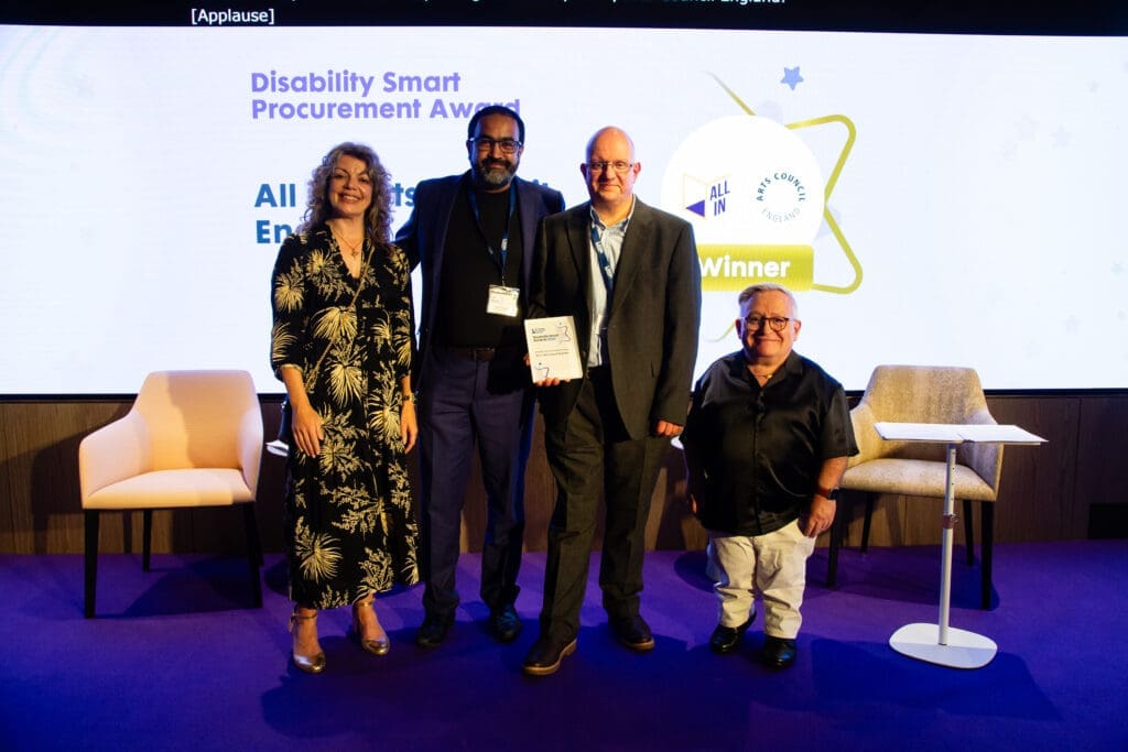 The All In, Arts Council England team posing on stage with ceremony hosts Diane Lightfoot (left) and Simon Minty (right) after winning the Procurement Award at the Disability Smart Awards 2025.