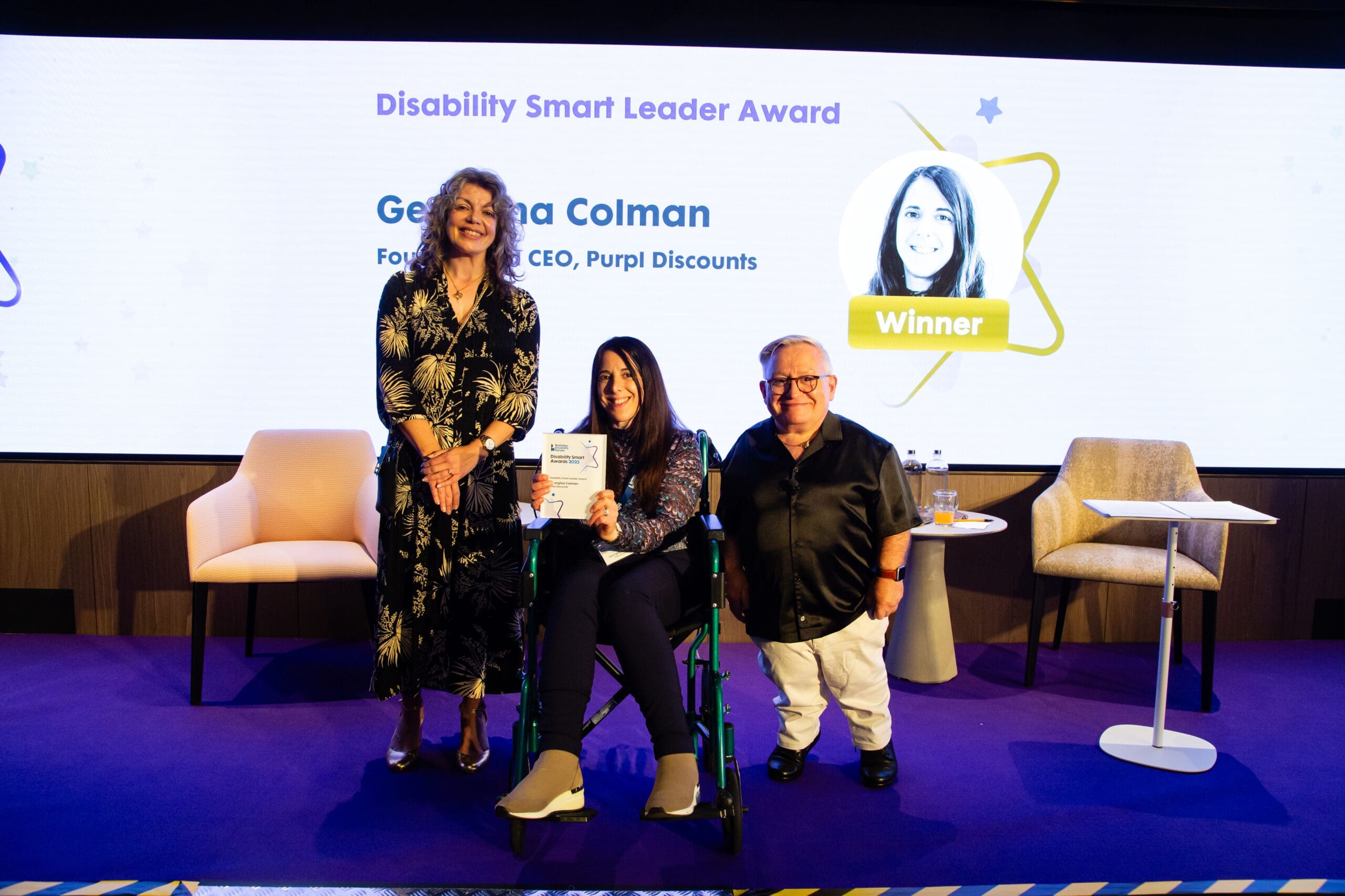 Georgina Colman receiving the Disability Smart Leader Award at the 2025 ceremony, seated at centre holding the award, with two presenters standing beside her on stage.