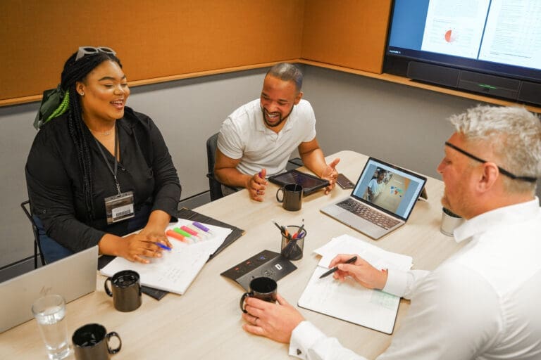 Photo of three colleagues in a meeting room at work with notebooks, pens and a laptop on the table