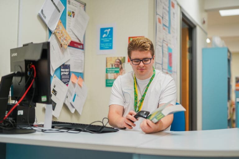 Library staff member scanning a book at a desk, wearing a sunflower lanyard that indicates hidden disabilities support.