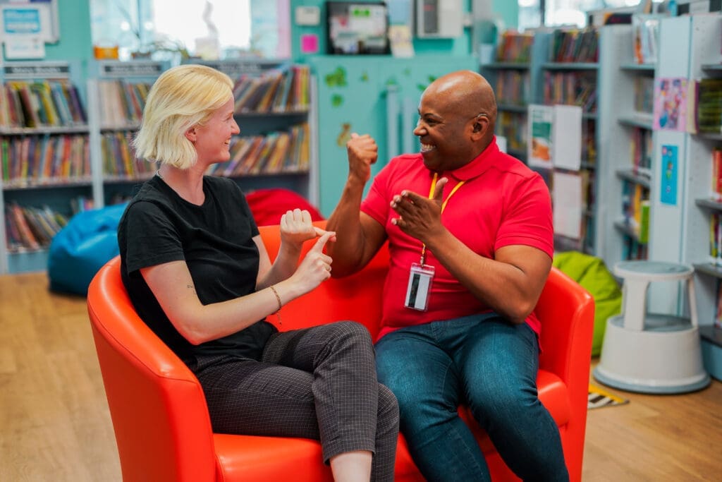 Two people sitting in bright orange chairs at a library, smiling and using sign language to communicate.