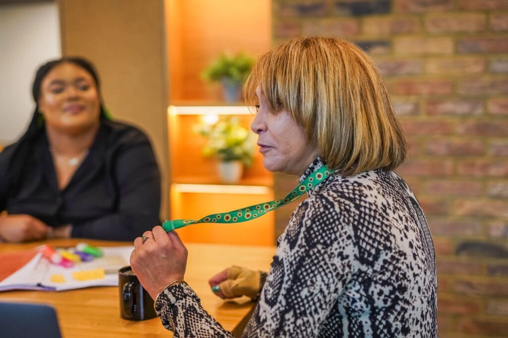 A woman wearing a sunflower lanyard sits at a table during a meeting, with another colleague blurred in the background.