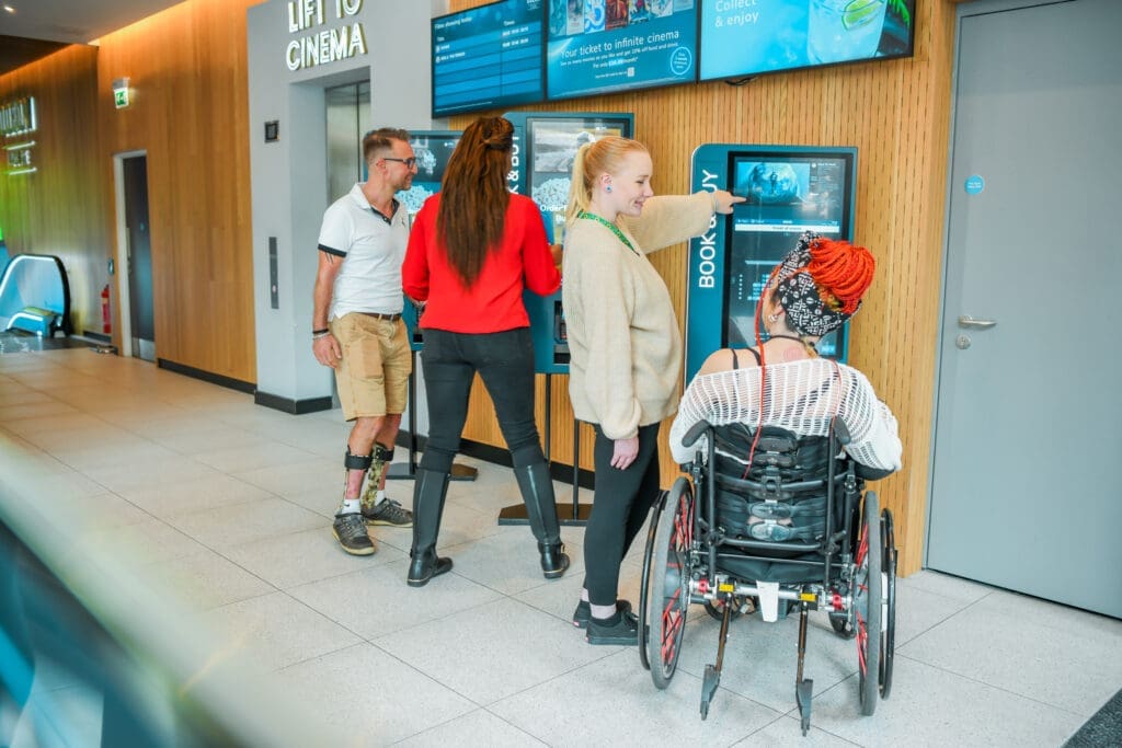 A group of people using self-service booking screens in a cinema lobby, including a wheelchair user and a man with a prosthetic leg.