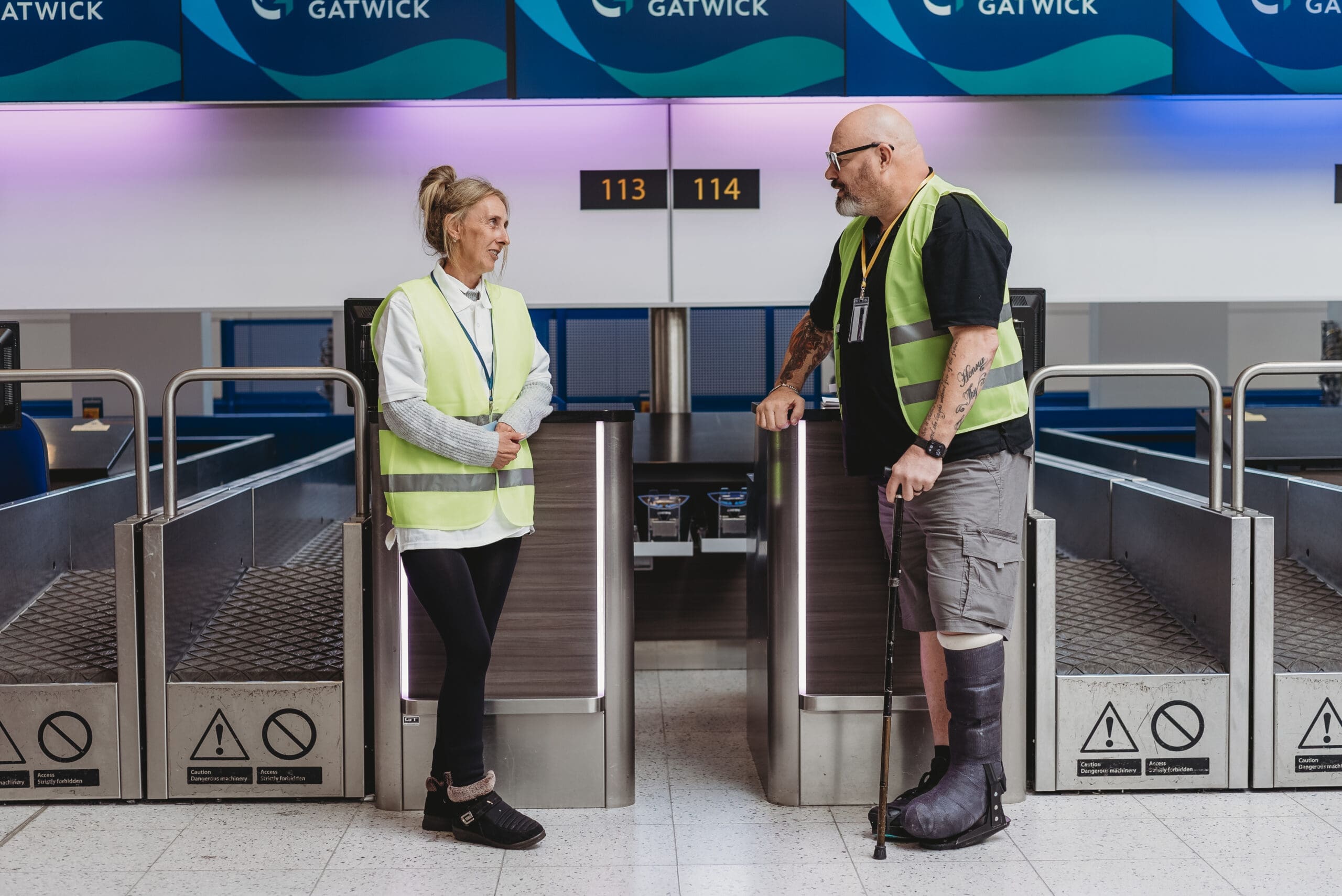 An airport worker who wears a medical boot and uses a walking stick talks to a colleague with less-visible disabilities