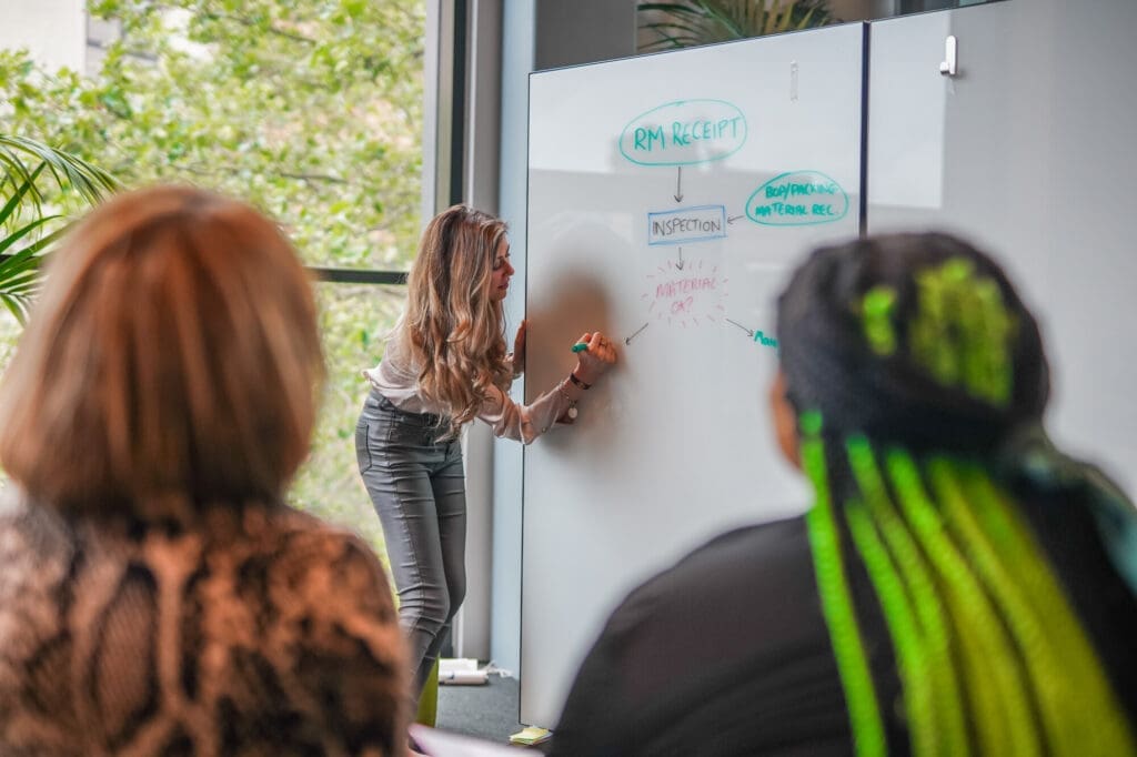 A person writes on a whiteboard during a training session, with two participants watching in the foreground.
