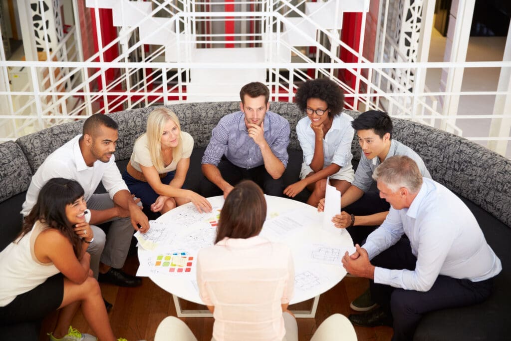 Group of eight colleagues sitting around a round table in a modern office, engaged in a collaborative discussion with papers and charts spread out.