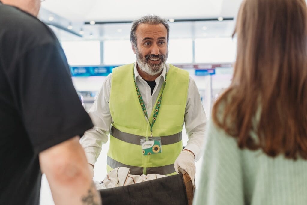 Person wearing a sunflower lanyard and high-visibility vest smiles while speaking with two passengers in an airport setting.
