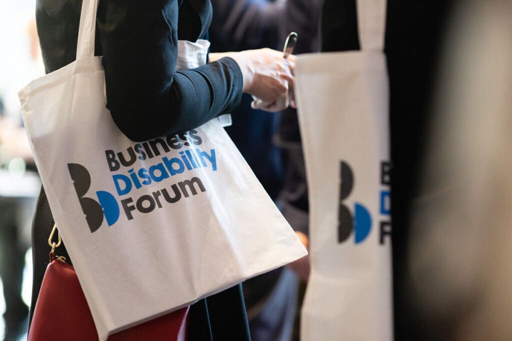 Close-up of attendees at an event holding white tote bags with the Business Disability Forum logo printed in black and blue.