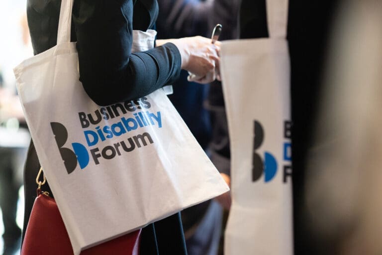 Close-up of attendees at an event holding white tote bags with the Business Disability Forum logo printed in black and blue.
