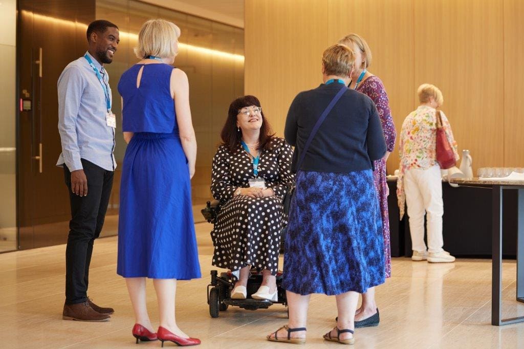 Group of 5 smiling delegates chatting at an event. In the centre is our Lead Business Partner, Rebecca Elliott, who uses a wheelchair.