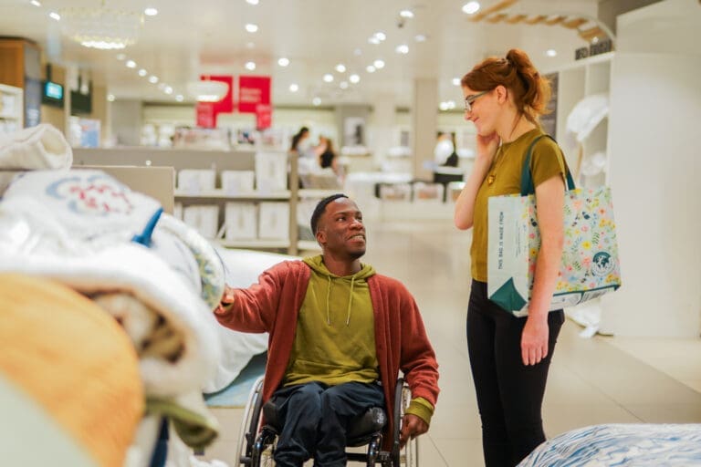 A ​wheelchair ​user ​with ​Spina ​Bifida ​browses ​soft ​furnishings ​in ​a ​store ​with ​a ​friend ​who ​has ​a ​less-​visible ​disability ​(​PMDD)​