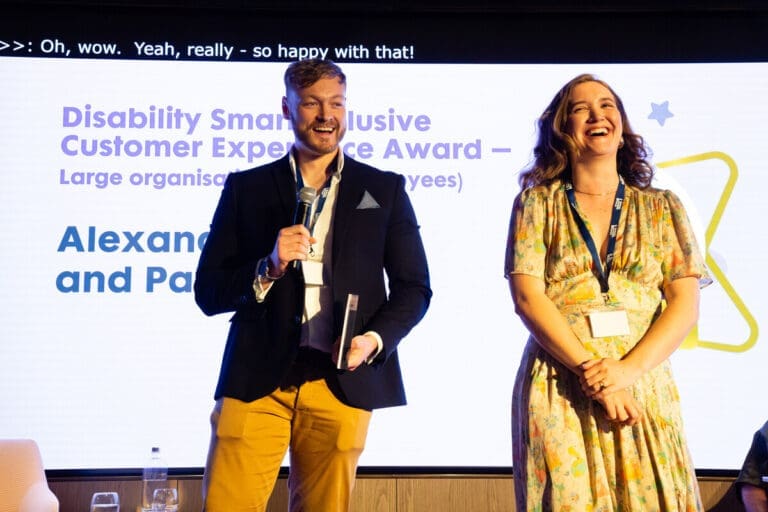 Two previous Disability Smart Impact Award winners laughing on stage. The person on the left is holding an award and speaking into a microphone.