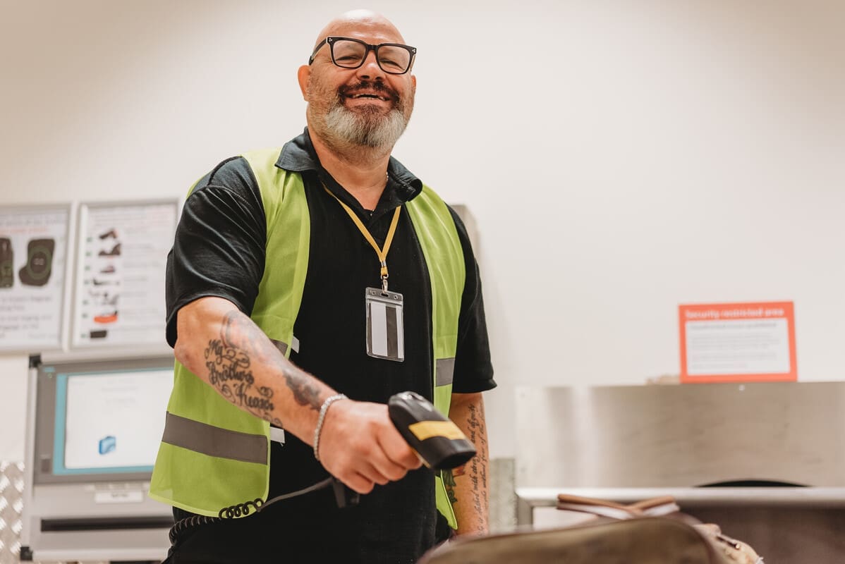 Person with a less-visible disability, who wears glasses an a high-vis jacket, smiles while holding a scanner in a warehouse.