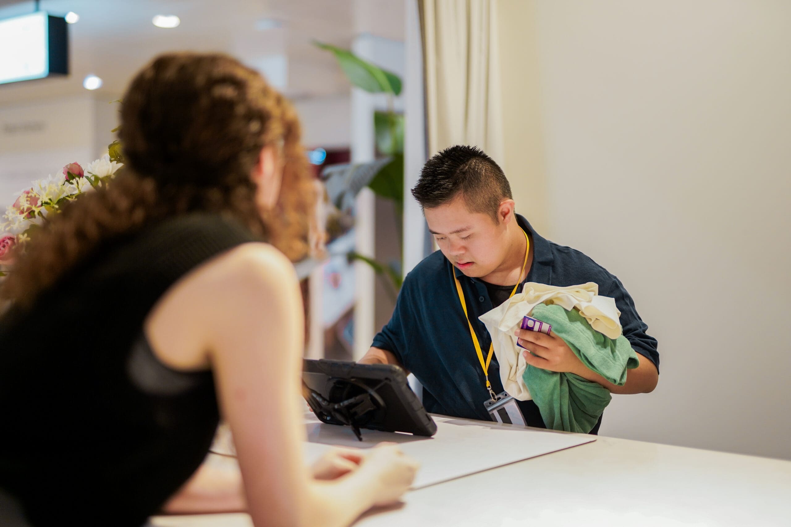 Retail employee with Down’s syndrome serving a customer at a checkout, holding folded clothing and using a point-of-sale tablet in a store setting