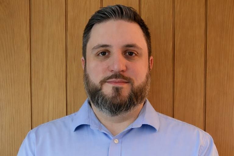 Man with short dark hair and a beard wearing a light blue button-down shirt, standing with hands together in front of a wooden panel background.