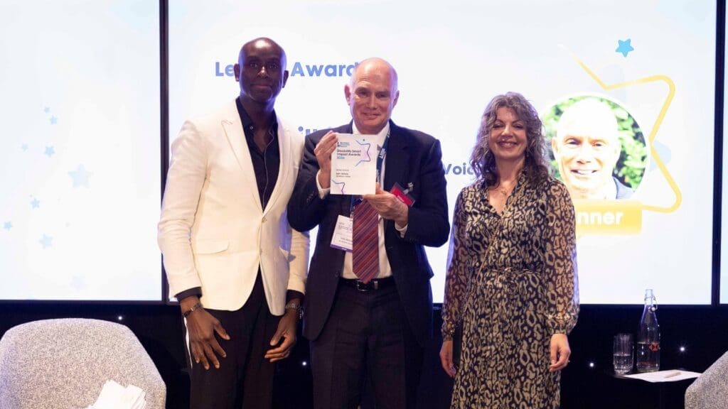 Iain Wilkie holding the Leader Award trophy on stage with presenters at the Disability Smart Impact Awards 2026.