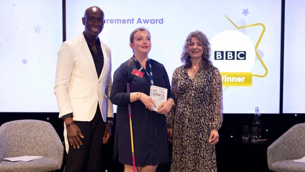 BBC representative holding Procurement Award on stage with presenters at the Disability Smart Impact Awards 2026.