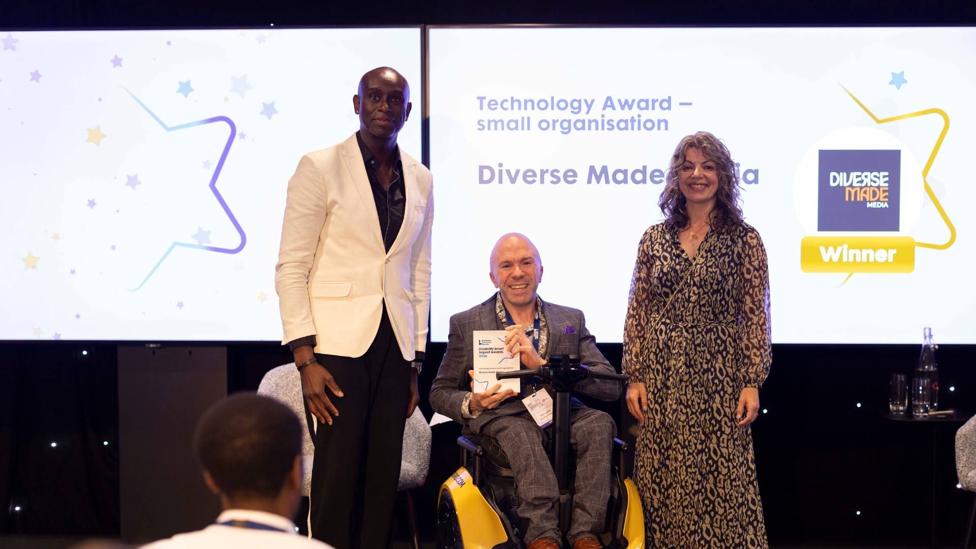 Diverse Made Media winner holding Technology Award while seated in a powered wheelchair on stage at the Disability Smart Impact Awards 2026.