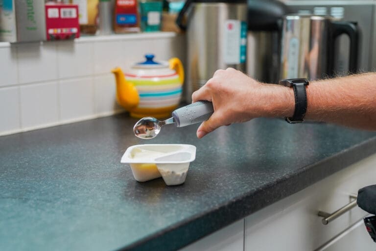 A person's hand grips an adapted spoon next to a pot of yoghurt